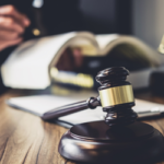 Gavel on a desk in the forefront with a person holding a law book and legal scales in the background