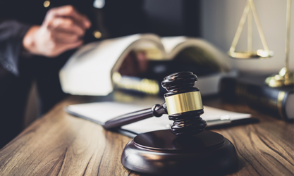 Gavel on a desk in the forefront with a person holding a law book and legal scales in the background