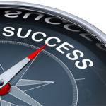 A dark gray compass with a chrome rim and a silver dial with a red tip pointed towards the word success