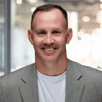 Light skinned man with very short brown hair wearing a white t-shirt with a neutral colored blazer