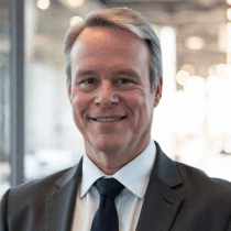 Man with light skin and short fair hair in a white shirt with a black tie and charcoal colored jacket.