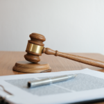 Gavel sitting on wooden table alongside a clipboard with paperwork with a pen sitting on top.