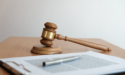 Gavel sitting on wooden table alongside a clipboard with paperwork with a pen sitting on top.