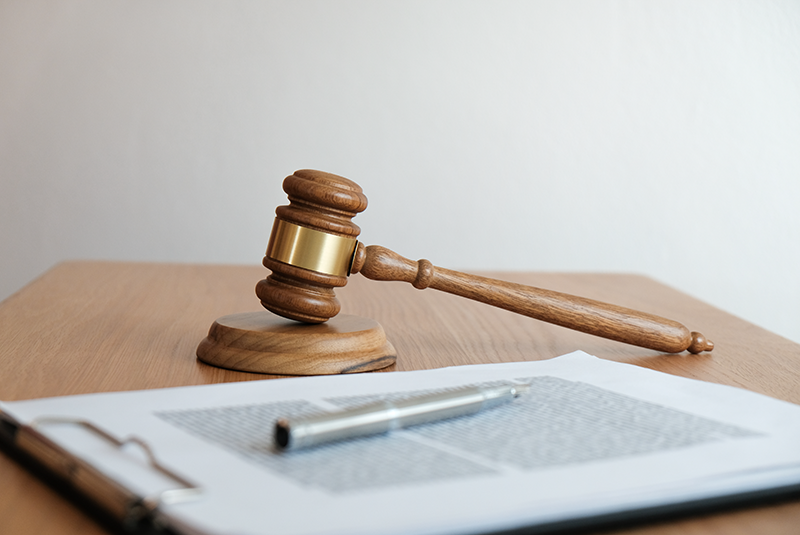 Gavel sitting on wooden table alongside a clipboard with paperwork with a pen sitting on top.