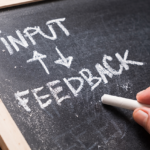 Dark chalkboard with a light wood border with the words input and feedback written in white chalk with directional arrows between the words.